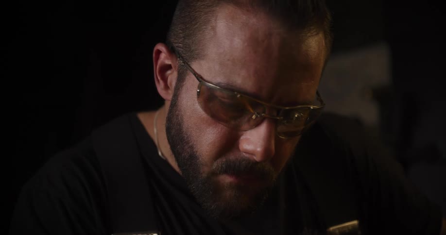 A focused craftsman wearing safety goggles concentrates intently on his work in a dimly lit workshop