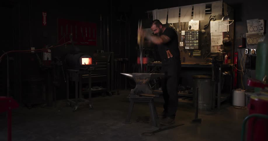 A bladesmith with tattoos hammers and shapes a glowing hot piece of steel on an anvil in a dark forge