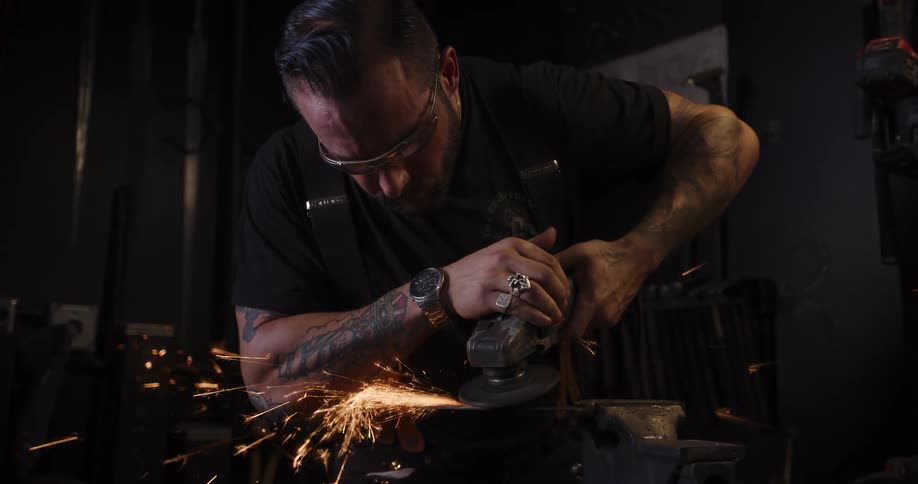 A tattooed metalworker in safety glasses uses an angle grinder on a steel blade, sending a cascade of bright sparks flying in slow motion