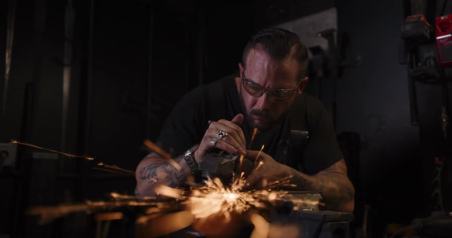 A tattooed craftsman meticulously grinds a piece of metal with an angle grinder, creating a spectacular shower of sparks in his dark workshop