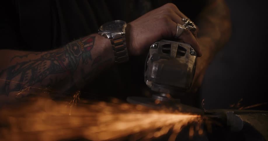 A close-up shot shows the tattooed hands of a craftsman using an angle grinder, sending a shower of glowing sparks flying