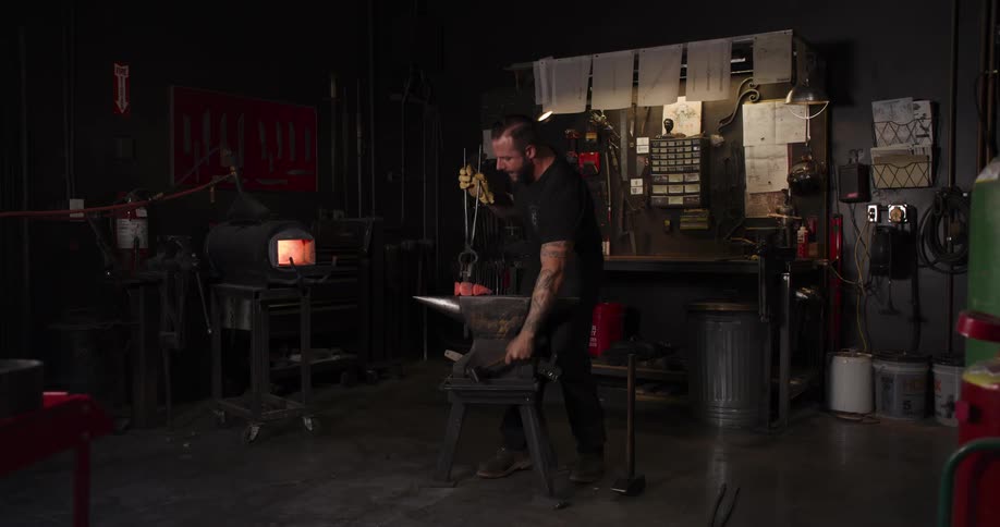 A male blacksmith forges a hot piece of glowing metal on an anvil in his workshop