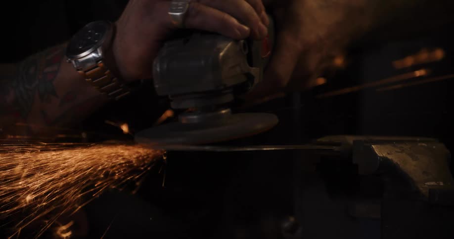 Slow motion close-up of a tattooed craftsman using an angle grinder on a metal blade, sending a shower of bright sparks flying in a dark workshop