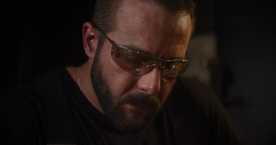 A close-up shot of a focused, bearded craftsman wearing safety glasses while concentrating on his work in a dimly lit workshop