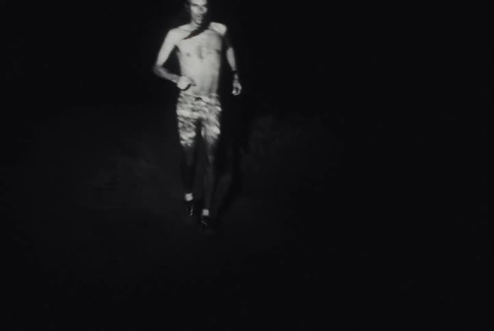 A gritty, black and white point-of-view shot follows a shirtless man running on a dark trail at night, illuminated only by his headlamp
