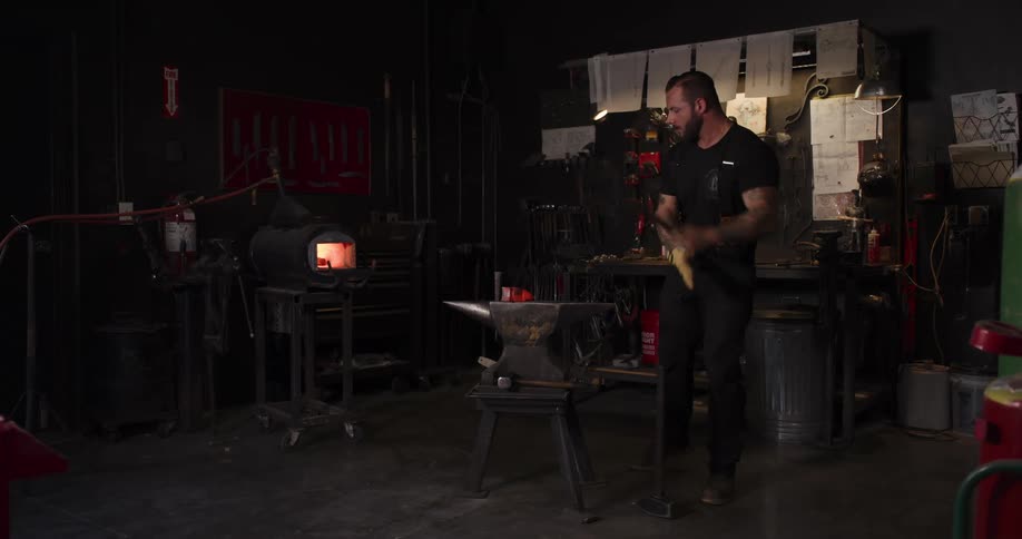 A strong, tattooed blacksmith hammers a glowing hot piece of metal on an anvil and returns another piece to the forge in his dark workshop