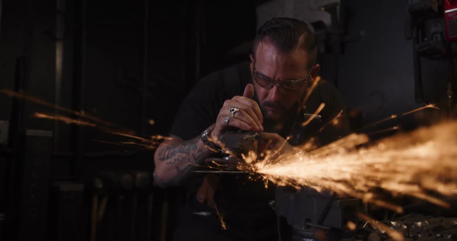 A tattooed metalworker in safety glasses uses an angle grinder, creating a brilliant shower of sparks in his dark workshop