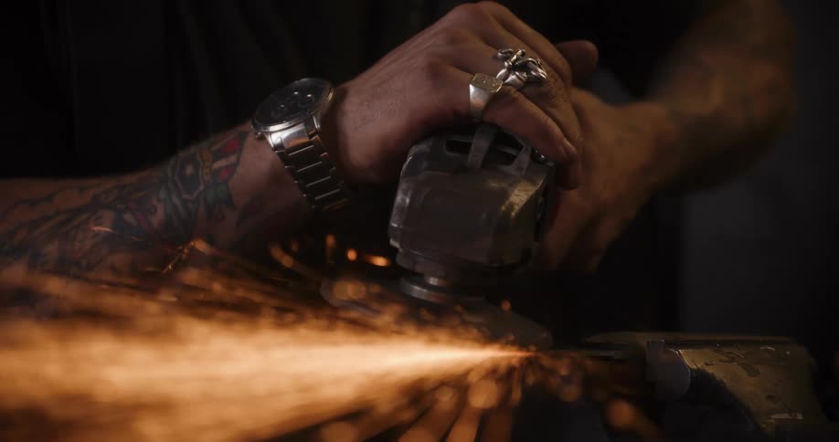 A tattooed craftsman uses an angle grinder, creating a shower of fiery sparks as he works on a piece of metal in a dark workshop