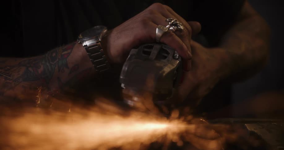Slow motion close-up of a tattooed metalworker's hands using an angle grinder, creating a spectacular shower of fiery orange sparks in a dark workshop