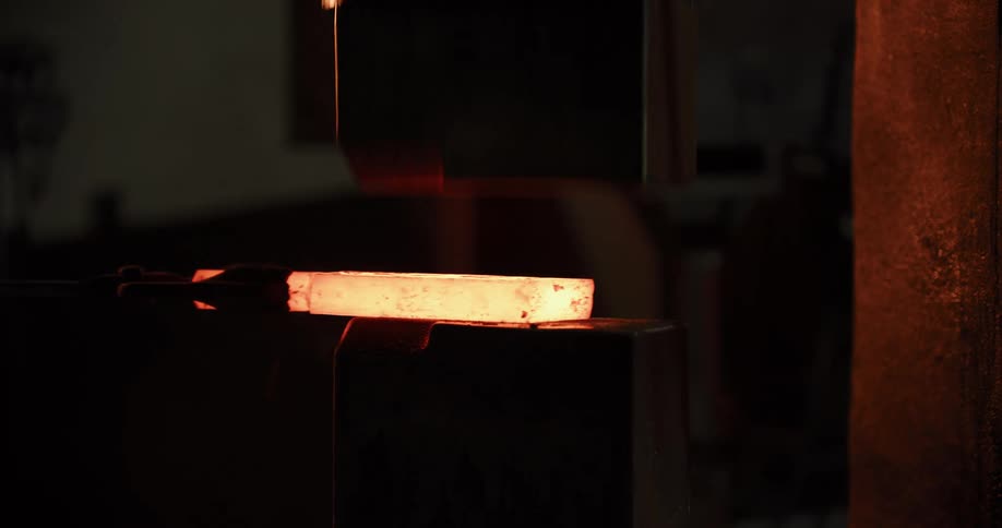 A hydraulic press flattens a glowing hot piece of metal held by tongs in a dark industrial workshop