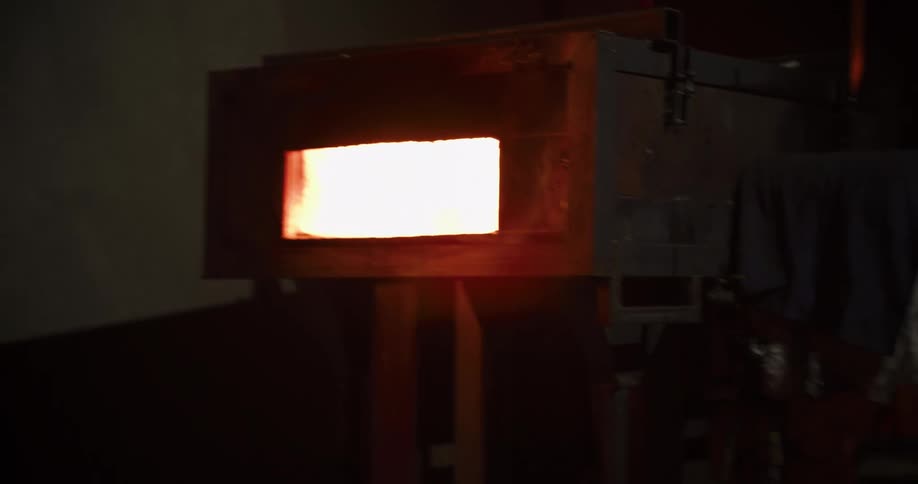 A metalworker removes a piece of glowing hot steel from a furnace in a dimly lit workshop in slow motion