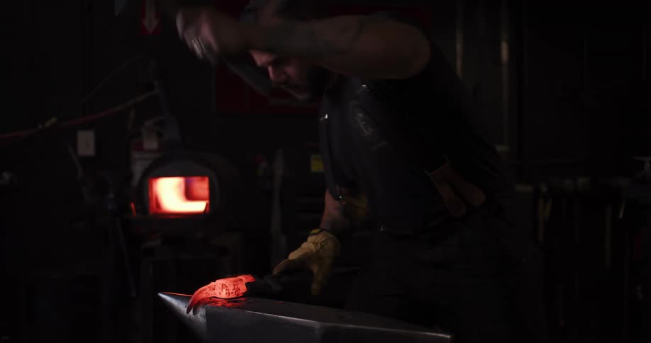 A focused blacksmith hammers a piece of glowing red-hot metal into shape on an anvil in his dark, industrial workshop
