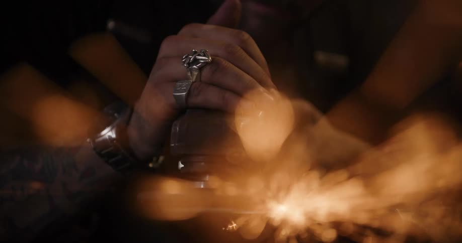 Dreamy slow-motion close-up of a tattooed man's hands grinding metal, creating a brilliant shower of sparks with an artistic bokeh effect