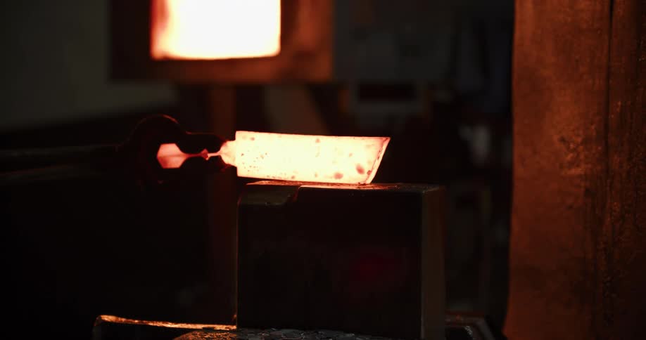 A blacksmith holds a glowing hot piece of molten metal with tongs in a dark workshop
