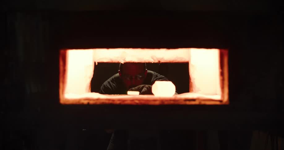 A craftsman wearing safety glasses carefully arranges glowing hot glass inside a fiery furnace