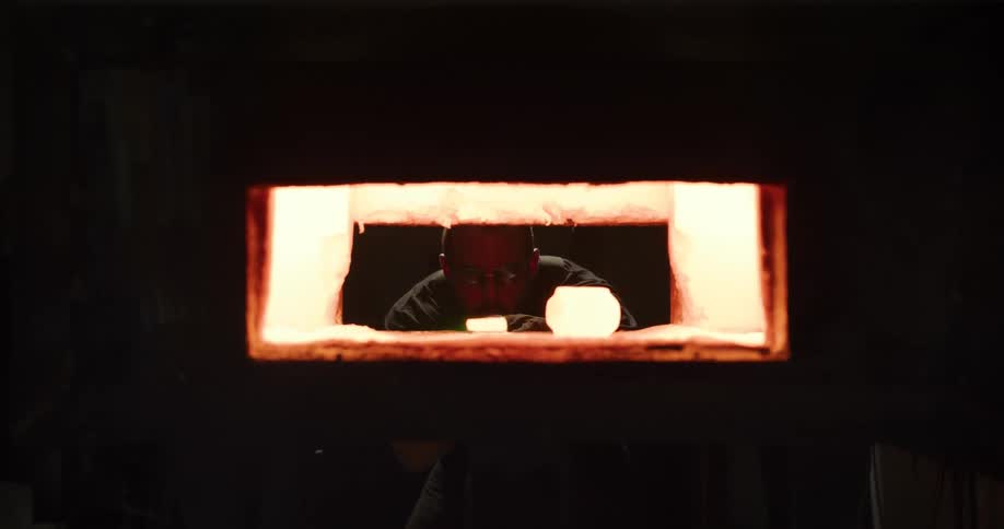 A glassblower wearing protective gear reaches into a hot kiln to remove a glowing piece of molten glass