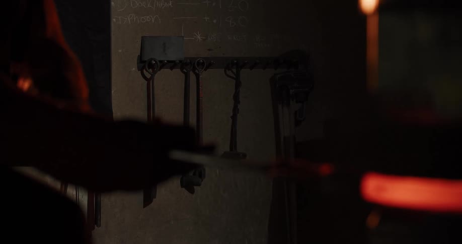 A rack of metal tools hangs on the wall of a dark workshop as a craftsman shapes a glowing hot piece of molten glass