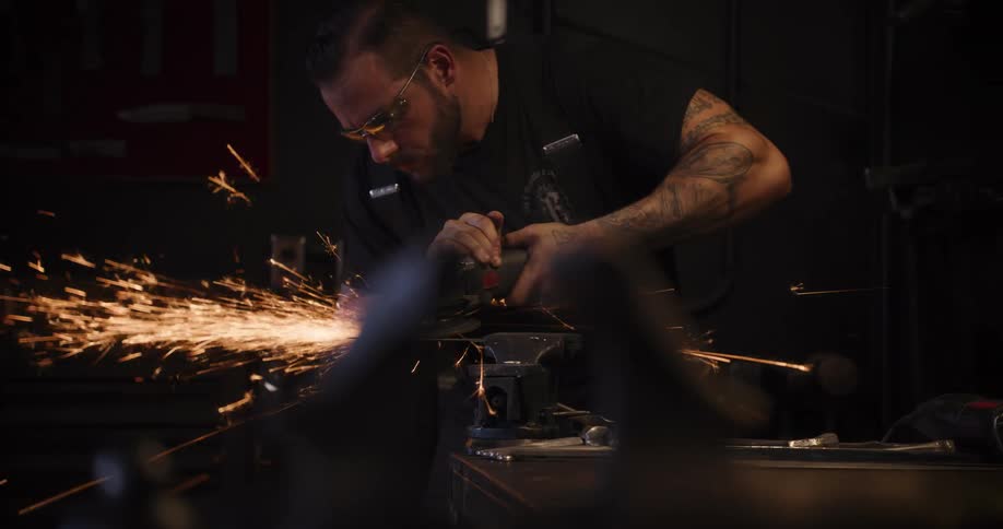 A tattooed blacksmith in safety glasses uses an angle grinder on a piece of metal, creating a brilliant shower of sparks in his workshop in slow motion