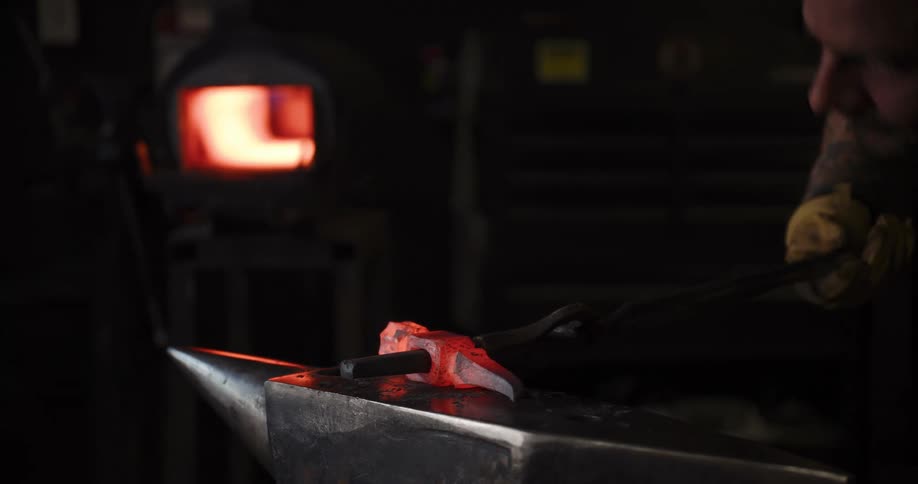 A skilled blacksmith hammers and shapes a glowing piece of red-hot metal on an anvil in a dark workshop