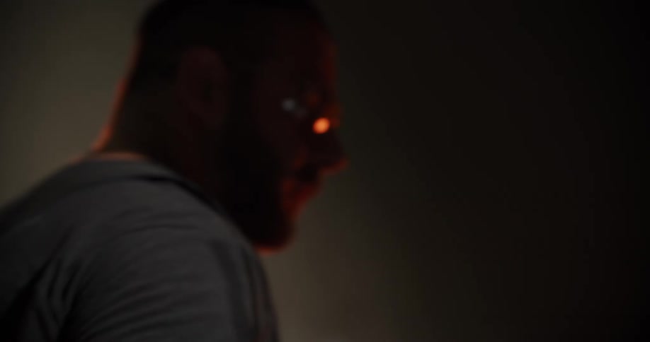 A dramatic, out-of-focus pan reveals the silhouette of a bearded man in a dark, industrial workshop
