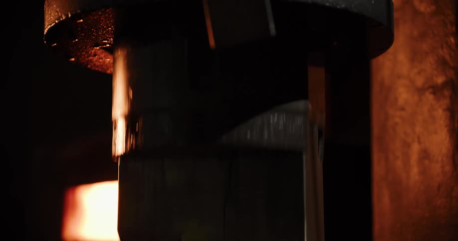 A powerful industrial pneumatic press repeatedly hammers a glowing red-hot metal bar in a dark forge