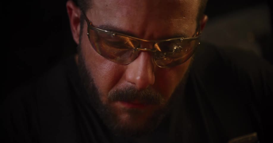 An intense close-up shot shows a focused craftsman wearing safety glasses with the warm glow of a fire reflected in the lenses