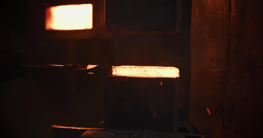 Slow motion shot of a hydraulic press forging a red-hot metal billet in a dark, industrial factory