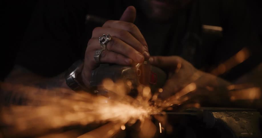 An intense close-up captures a tattooed metalworker's hands using an angle grinder, creating a shower of glowing sparks with a dreamy lens flare effect