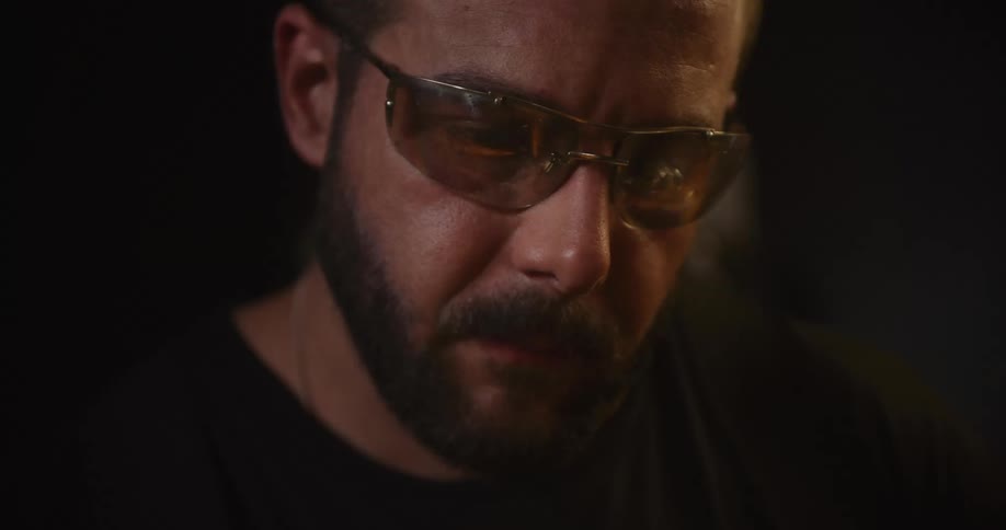 Close-up of a bearded artisan in safety glasses, his face illuminated by the flickering warm light of a fire in his dark workshop