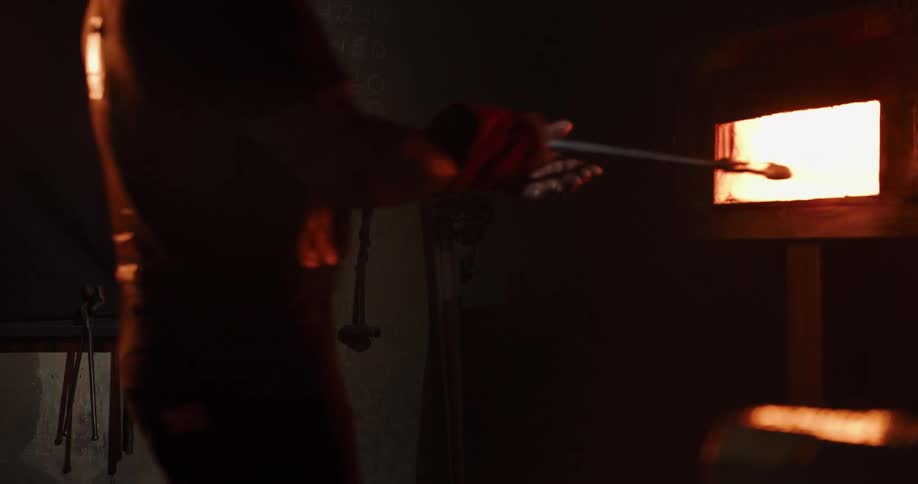A craftsman wearing protective gloves carefully heats a metal rod in a blazing furnace inside a dark workshop