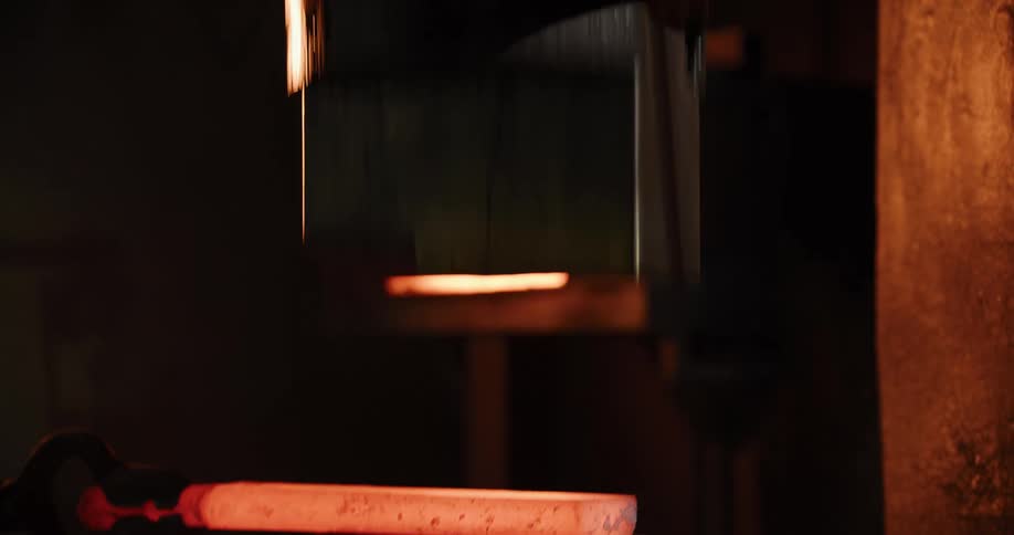 A powerful industrial press repeatedly hammers and forges a glowing piece of red-hot steel in a dark factory