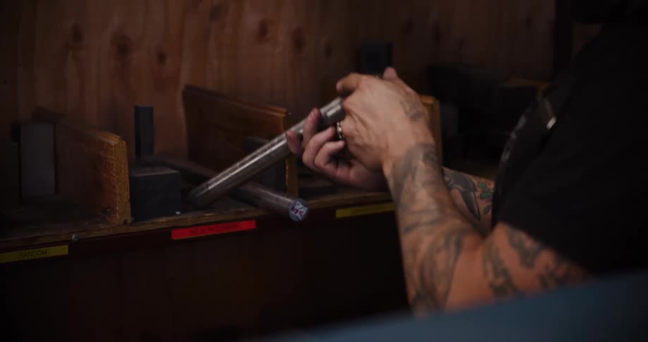 A tattooed worker inspects a metal rod and places it back on a wooden shelf in a workshop