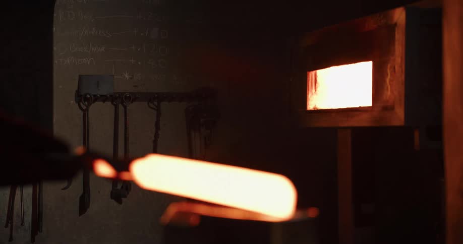 A blacksmith pulls a glowing piece of metal from a furnace and forges it with a hydraulic press in a dark workshop