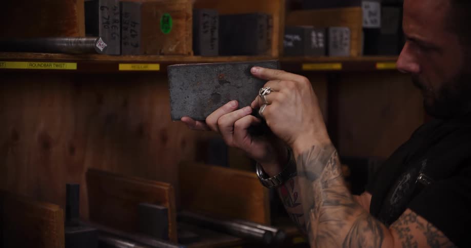 A tattooed metalworker carefully pulls a steel billet from a wooden shelf and inspects it in his workshop