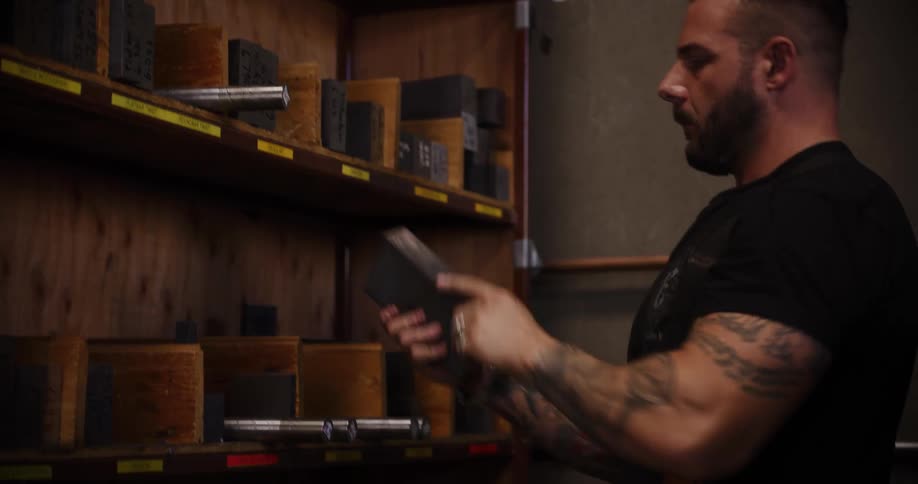A muscular, tattooed craftsman carefully selects a metal block from a wooden shelf in his industrial workshop