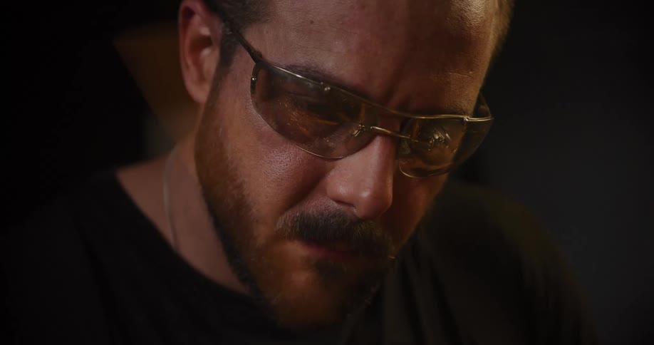 Close up shot of a focused bearded craftsman in safety glasses working intently as bright sparks fly in a dimly lit workshop