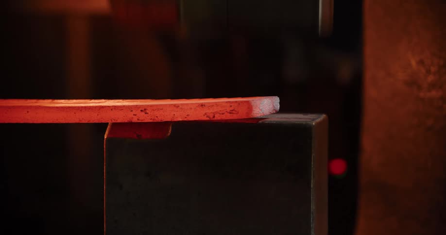 A red-hot metal billet is shaped and flattened by an industrial power hammer in a dark forge