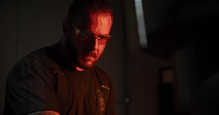 An intense, sweaty craftsman in safety glasses focuses intently on his work, illuminated by the red glow of a forge in his dark workshop