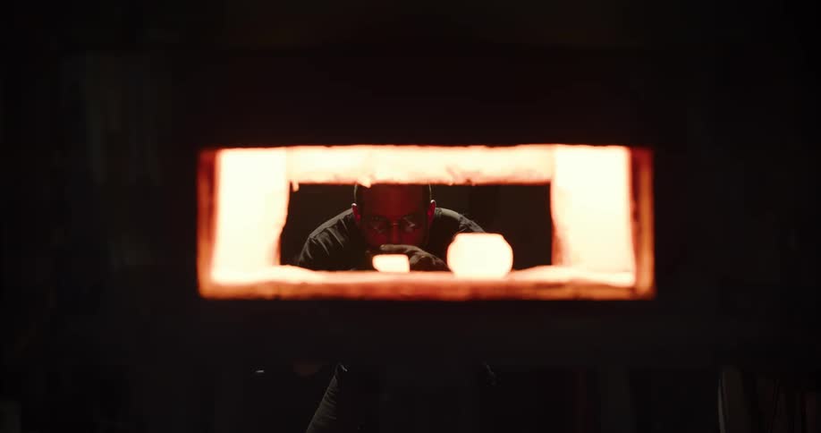A glassblower carefully places pieces of hot glass into a glowing furnace in his dark workshop