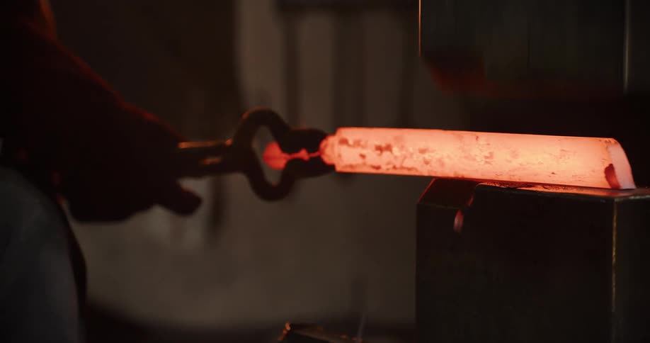 Close up slow motion shot of a blacksmith using tongs to hold and shape a red-hot metal billet under an industrial power press