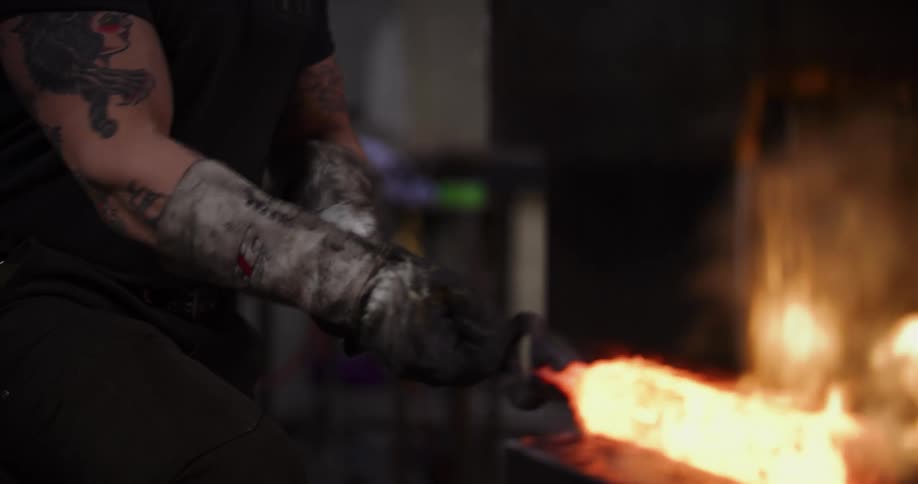 A close-up of a tattooed bladesmith wearing protective gloves while working with a glowing piece of hot metal in a forge