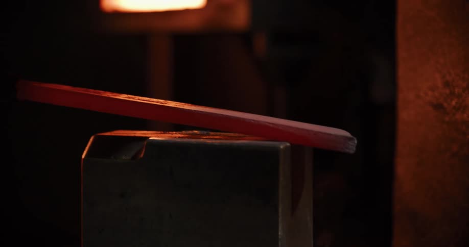 A powerful pneumatic hammer forges and flattens a piece of red-hot metal in a dark workshop