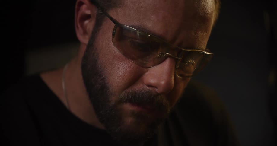 A focused craftsman in safety glasses works in a dark shop as a shower of bright sparks flies in the foreground