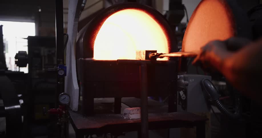 A blacksmith with tattooed arms heats a piece of metal in a glowing hot forge inside a dark workshop