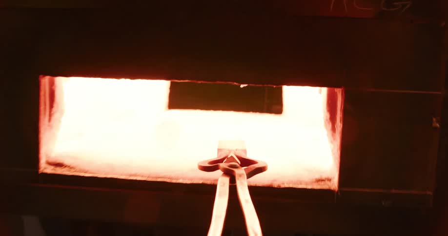 POV of a blacksmith carrying a hot block of steel with tongs from a power hammer into a fiery furnace