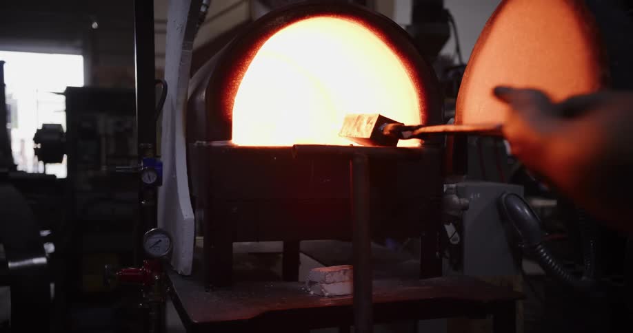 A craftsman with tattooed arms uses tongs to place a hot piece of metal into a glowing furnace in a dark workshop
