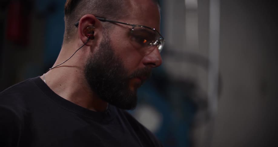 A bearded industrial worker wearing safety glasses and earplugs concentrates intently on his task in a workshop