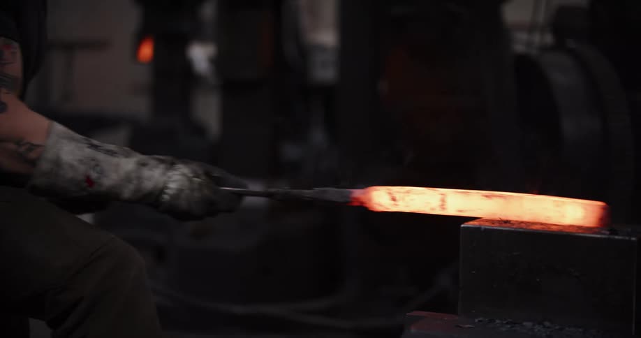 A blacksmith with tattooed arms uses tongs to position a glowing, red-hot metal billet on an anvil in a dark forge