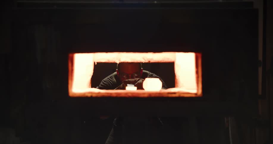 A glassblower opens a hot, glowing furnace door, carefully removes a piece of molten glass on a blowpipe, and then closes the door