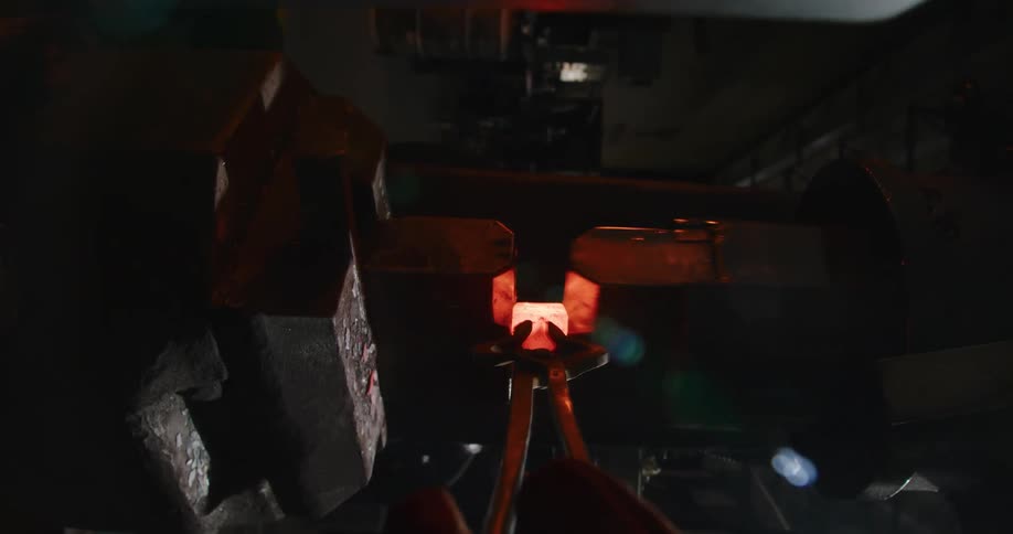 In slow motion, a power hammer strikes a glowing piece of metal held in tongs, sending a shower of sparks flying in a dark forge